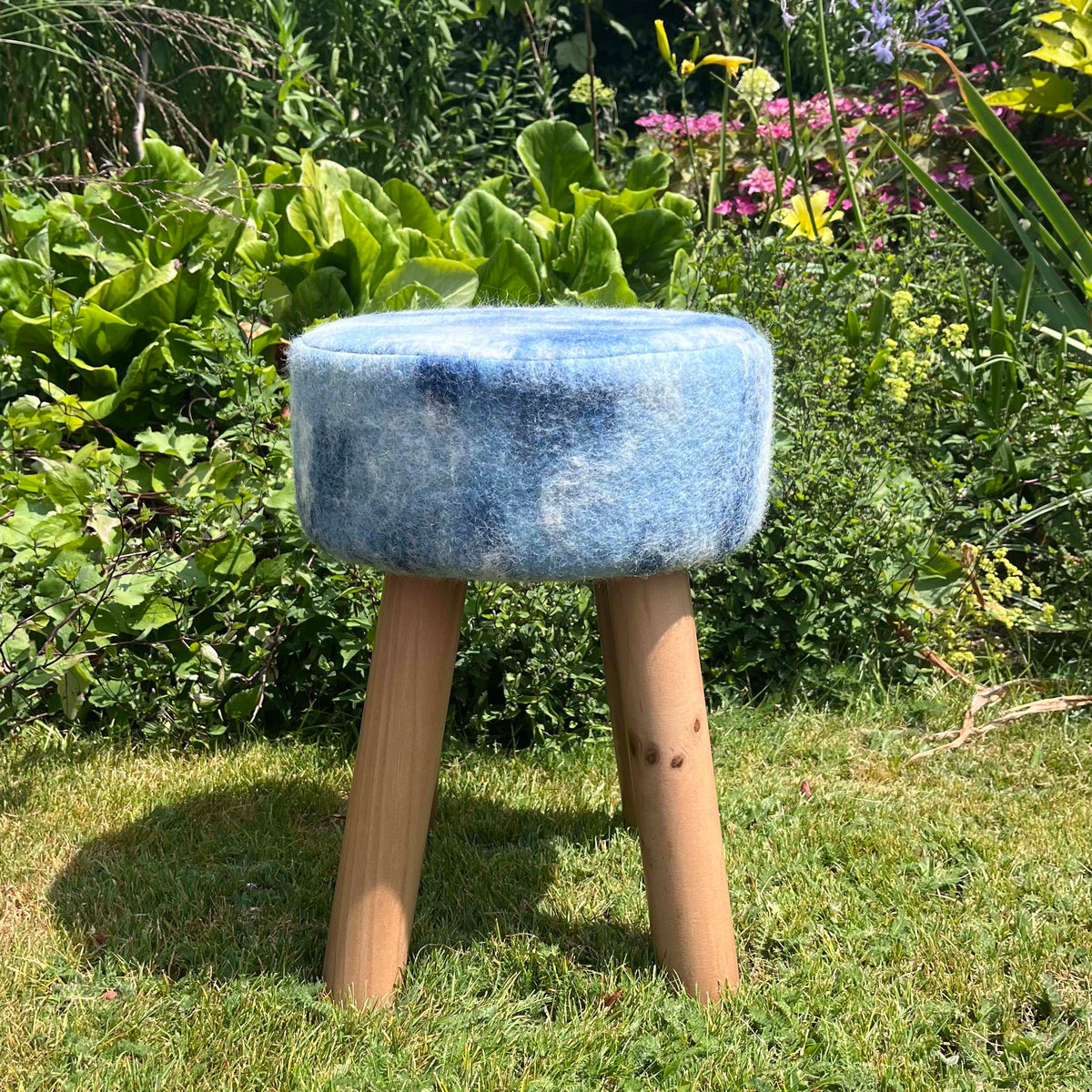 A small round stool with a blue upholstered seat made from felted British wool and four natural wood legs, photographed outdoors on grass with garden plants in the background. 