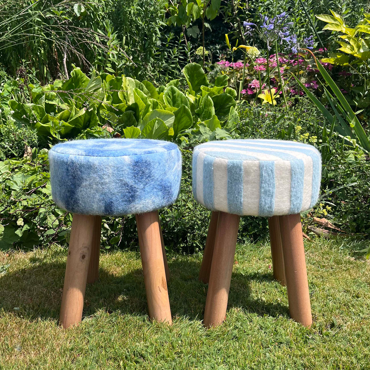 Two round upholstered stools with wooden legs placed on grass in a garden. The left stool has a blue wool seat, and the right stool has light blue and white vertical stripes, also made from felted British wool. 