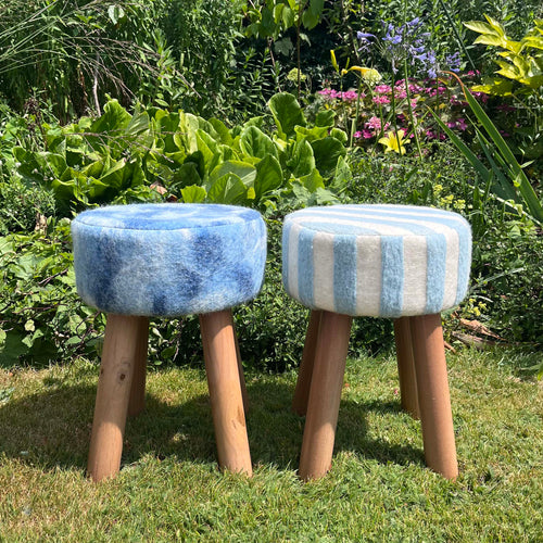 Two round upholstered stools with wooden legs placed on grass in a garden. The left stool has a blue wool seat, and the right stool has light blue and white vertical stripes, also made from felted British wool. 